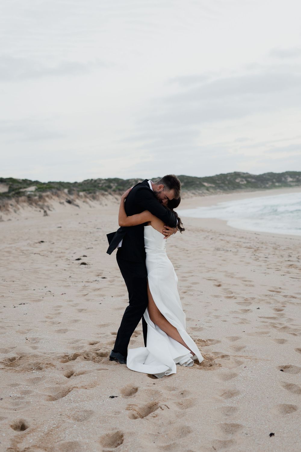 Intimate embrace between newlyweds on a sandy beach with ocean waves in the background.