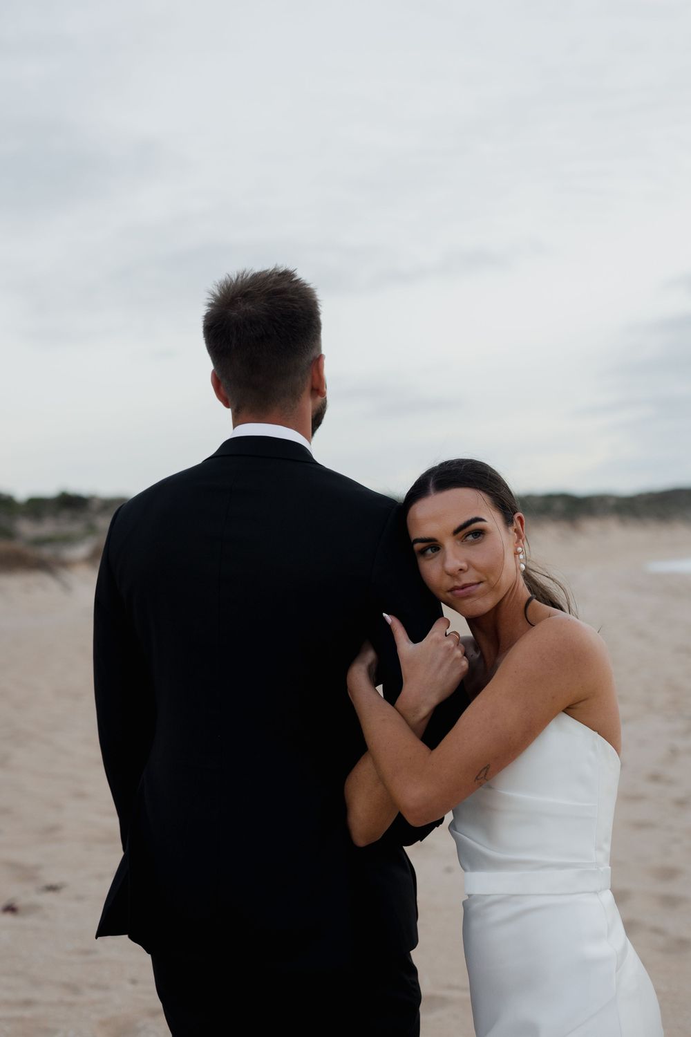 Intimate portrait of couple in wedding attire embracing on beach with overcast sky.