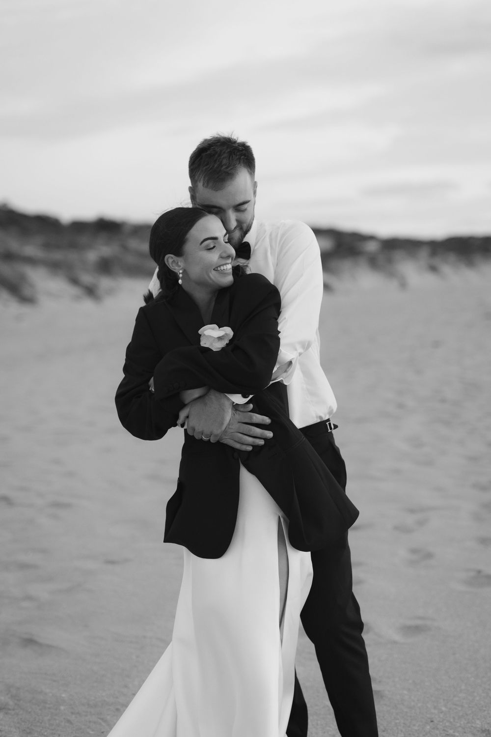 A romantic black and white portrait of newlyweds embracing on a beach.