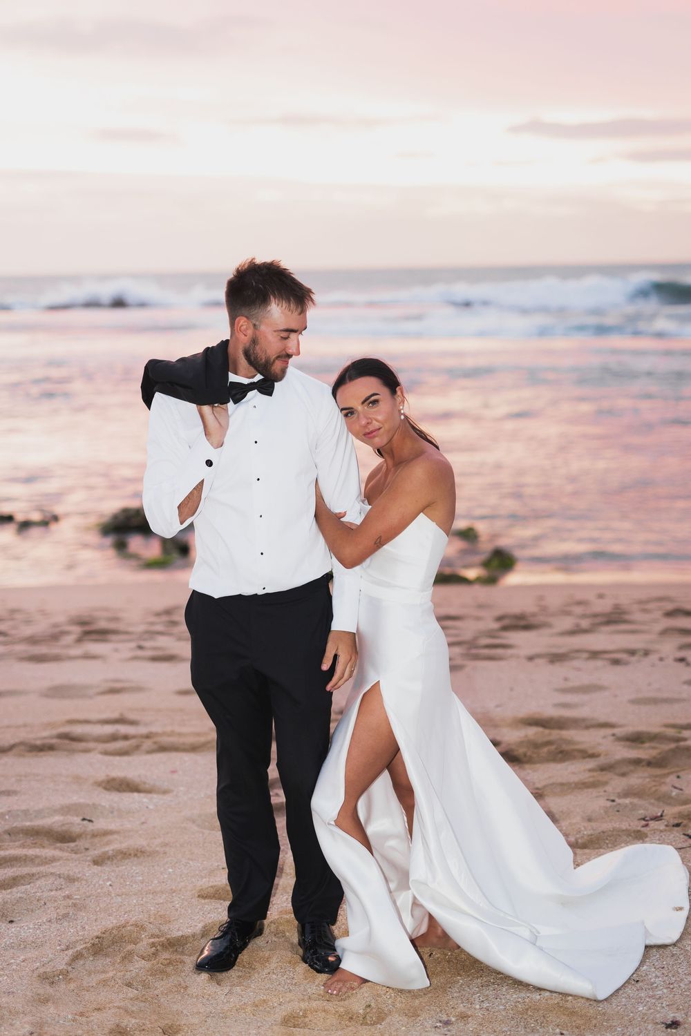 Newlyweds share a playful moment on a beach during sunset in formal attire.