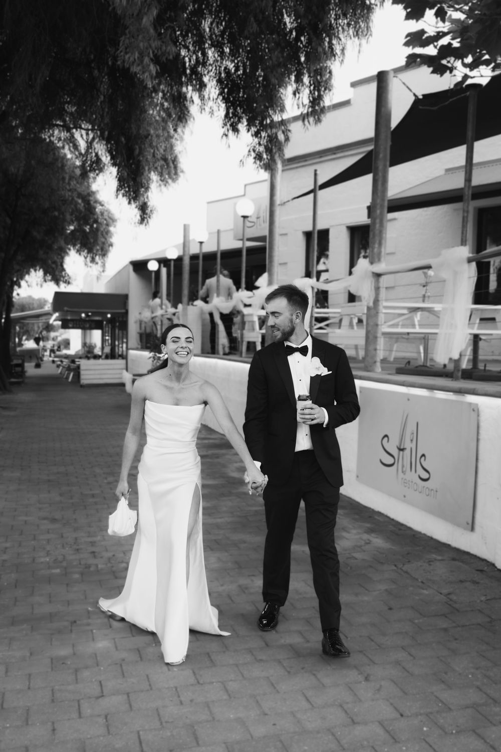 A couple walks together on a city sidewalk in elegant wedding attire.
