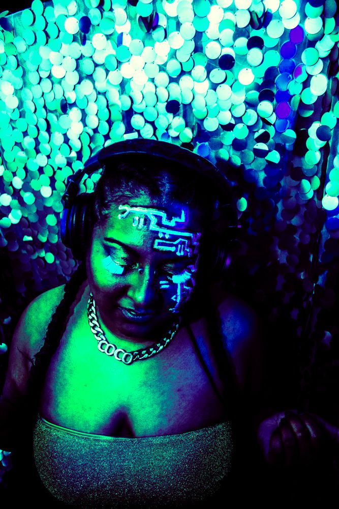 Neon-lit portrait with glowing turquoise bokeh background showing dramatic cyberpunk-style face paint and metallic necklace.