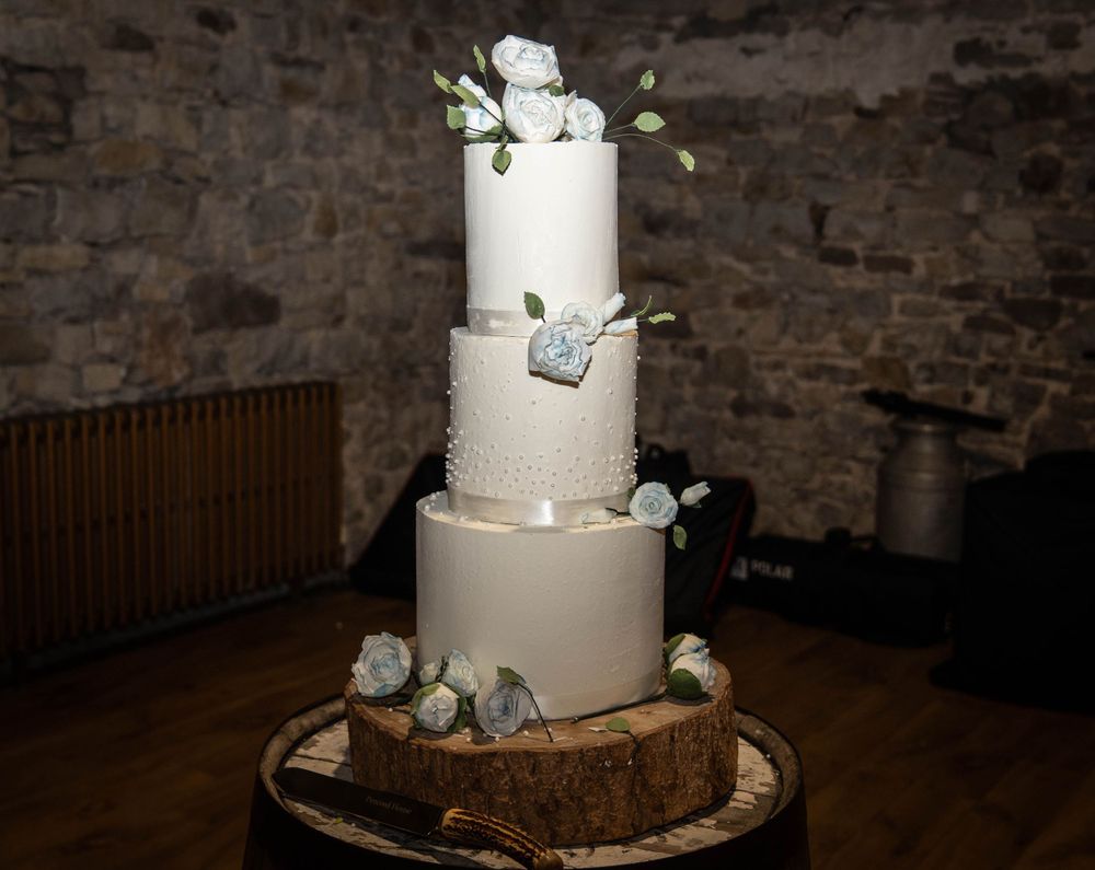 White wedding cake with floral decorations displayed on wooden base against stone wall background.
