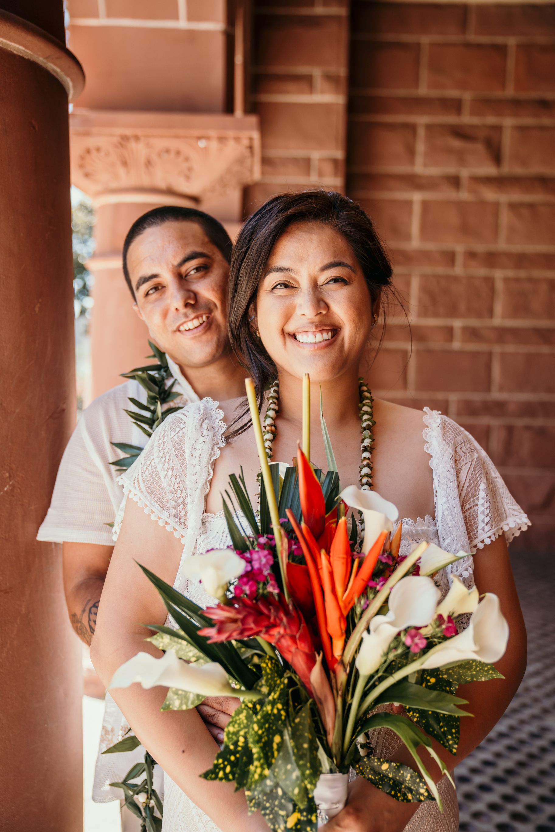 A happy couple poses together against a brick wall backdrop with a colorful tropical flower bouquet.