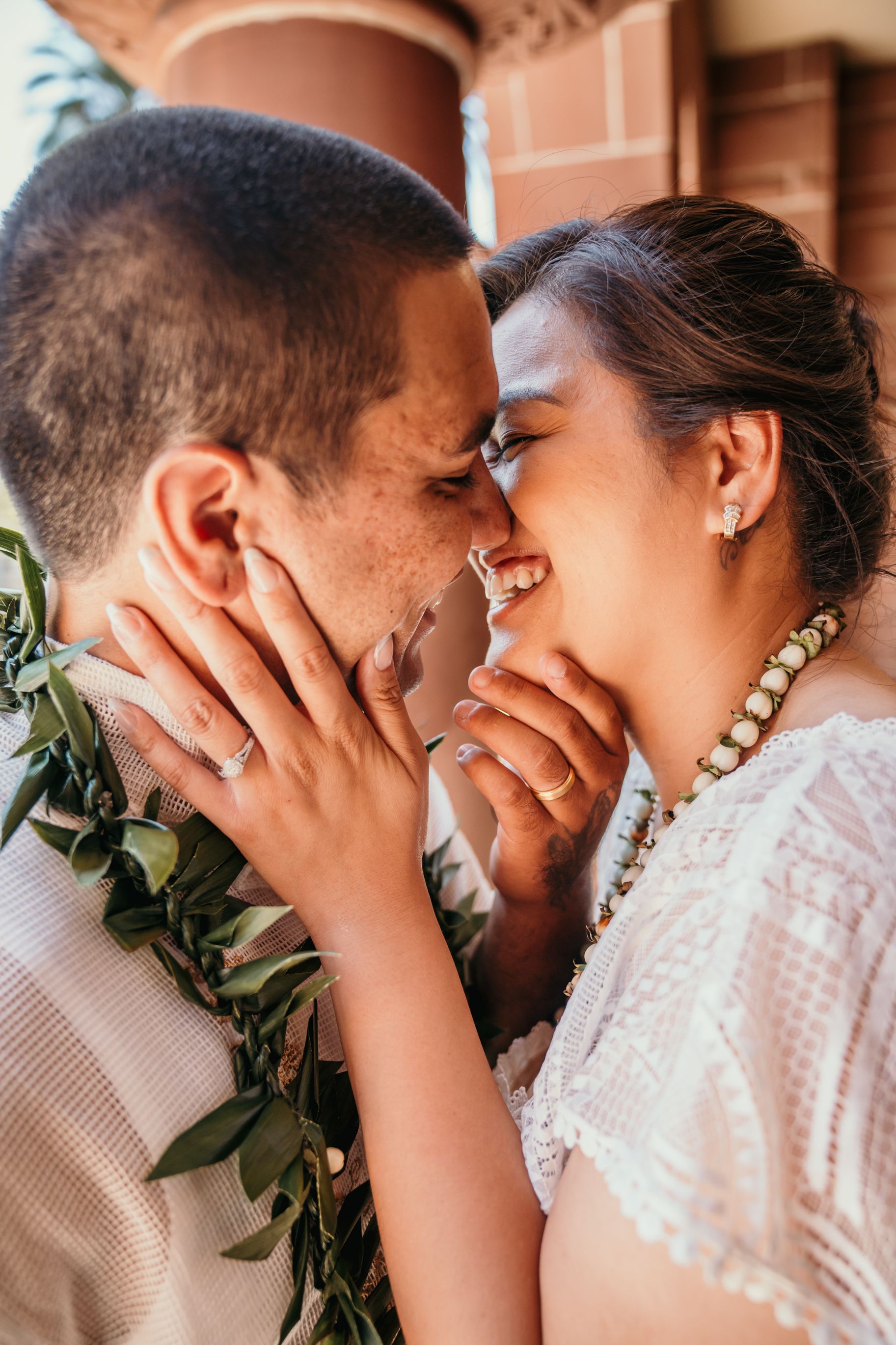 A romantic couple shares an intimate moment against a brick wall backdrop wearing traditional Hawaiian leis.