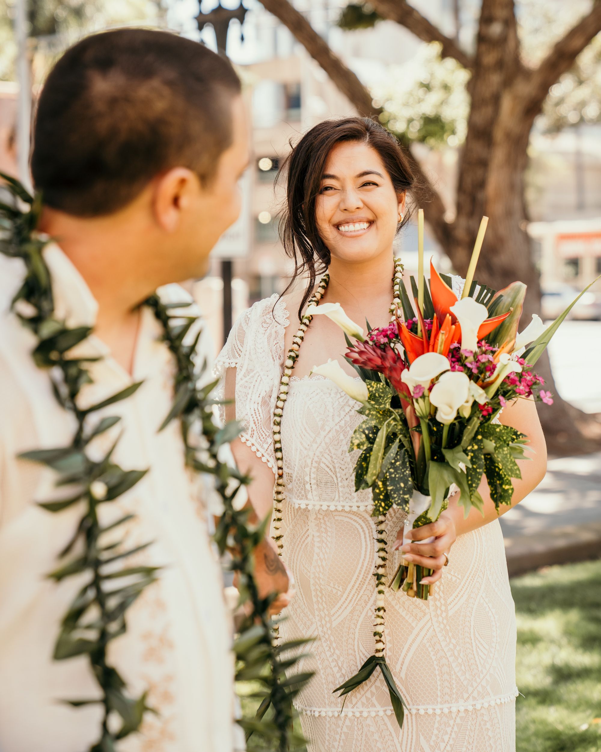 A couple shares a joyful moment outdoors, with one person holding a vibrant tropical bouquet of red and white flowers.