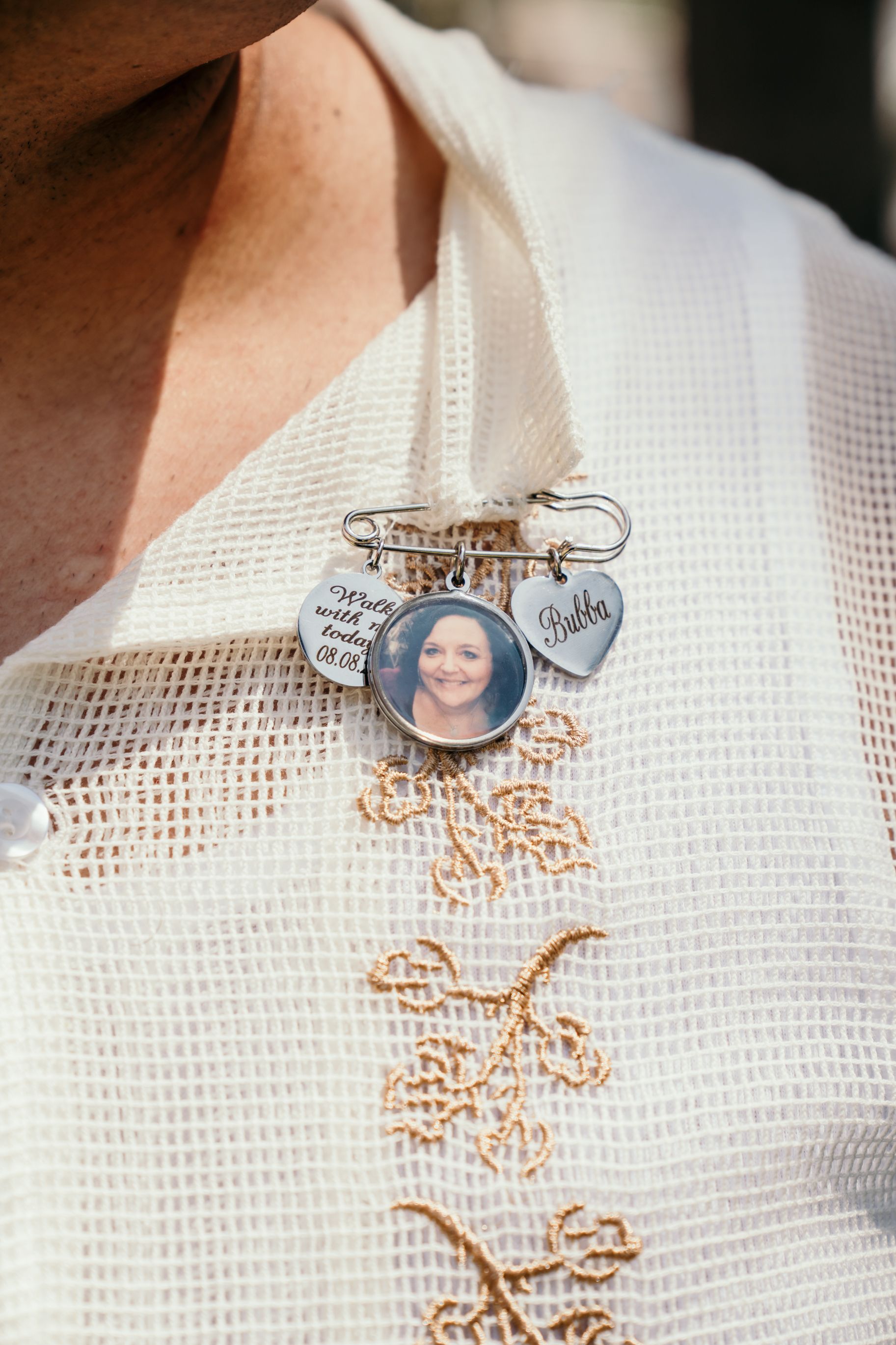 A memorial pin brooch with charms attached to white embroidered fabric shows delicate floral stitching details.