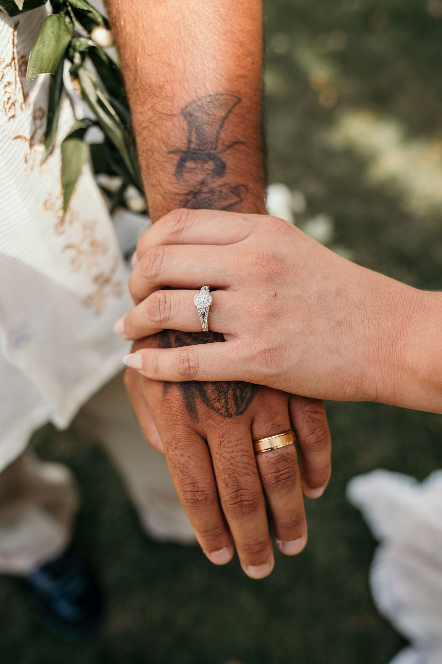 Two hands with wedding rings intertwined, one showing a visible tattoo against a green leafy background.
