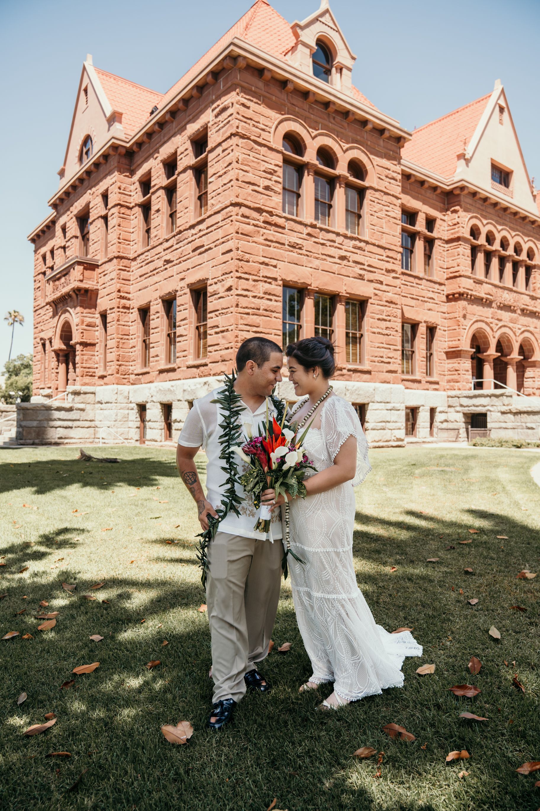A couple shares an intimate moment on the lawn of a grand historic brownstone building with Romanesque architecture.