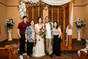 Wedding party poses beneath a draped arch adorned with white floral arrangements in an elegant indoor venue.