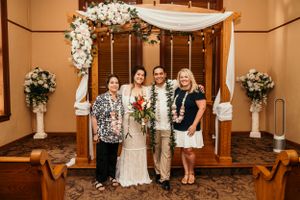 A wedding ceremony setup with a wooden arch decorated with white flowers and draping fabric in an indoor venue.