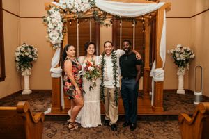 Wedding ceremony setup with wooden arch decorated with white florals and lights in an indoor venue.