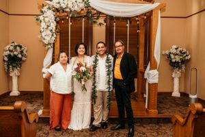 Wedding ceremony setup with white floral arch, draped fabric, and wedding party standing together indoors.