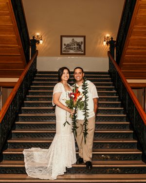 A couple in wedding attire poses on an elegant wooden staircase with wall sconces and a framed artwork in the background.