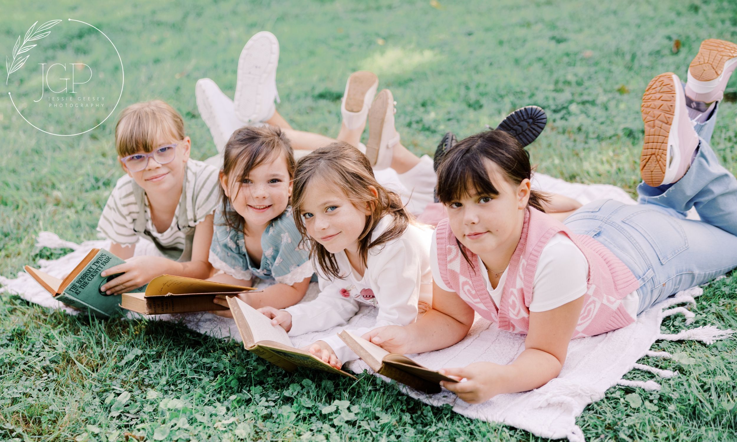 Four girls reading books on a blanket in green grass smiling into the camera