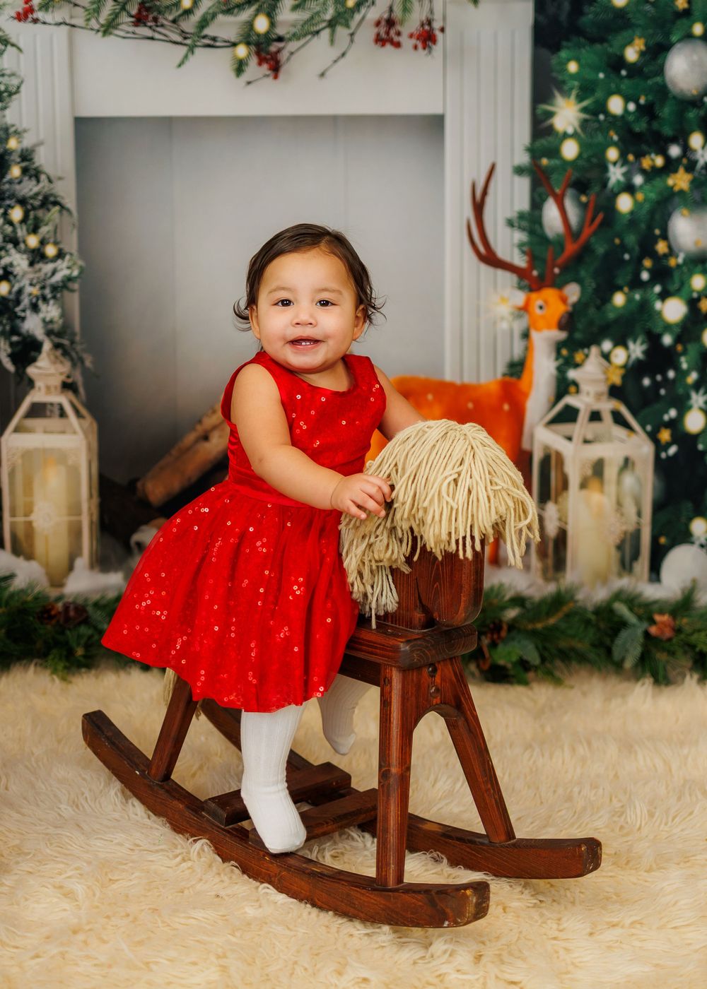 A baby in a red dress sits on a wooden rocking horse near a decorated Christmas tree and holiday decor.