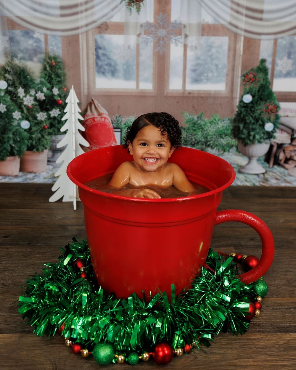 A child sits in a large red coffee cup prop surrounded by green tinsel garland and holiday decorations.