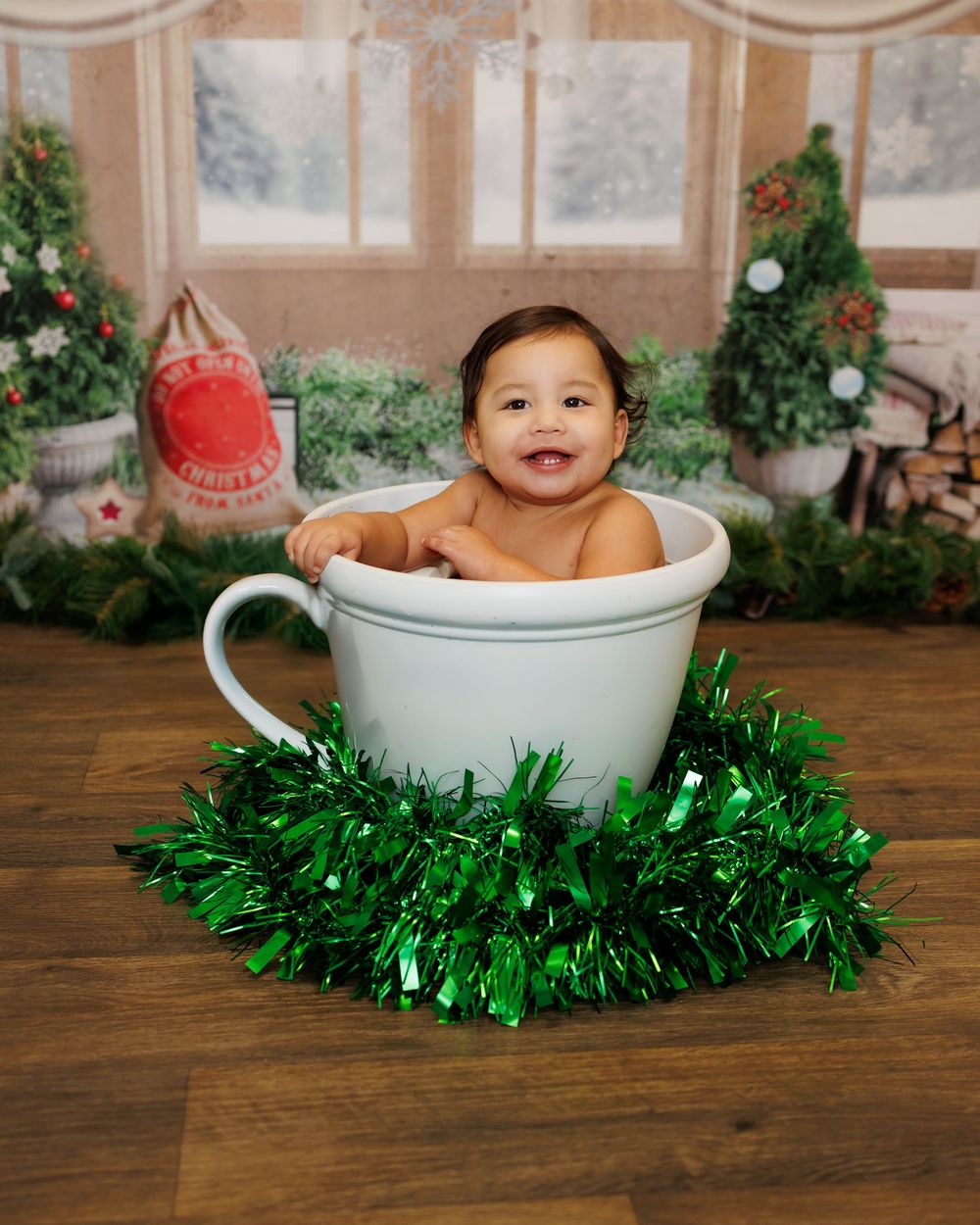 A baby sits in a white ceramic bowl surrounded by sparkly green Christmas tinsel garland on a wooden surface.
