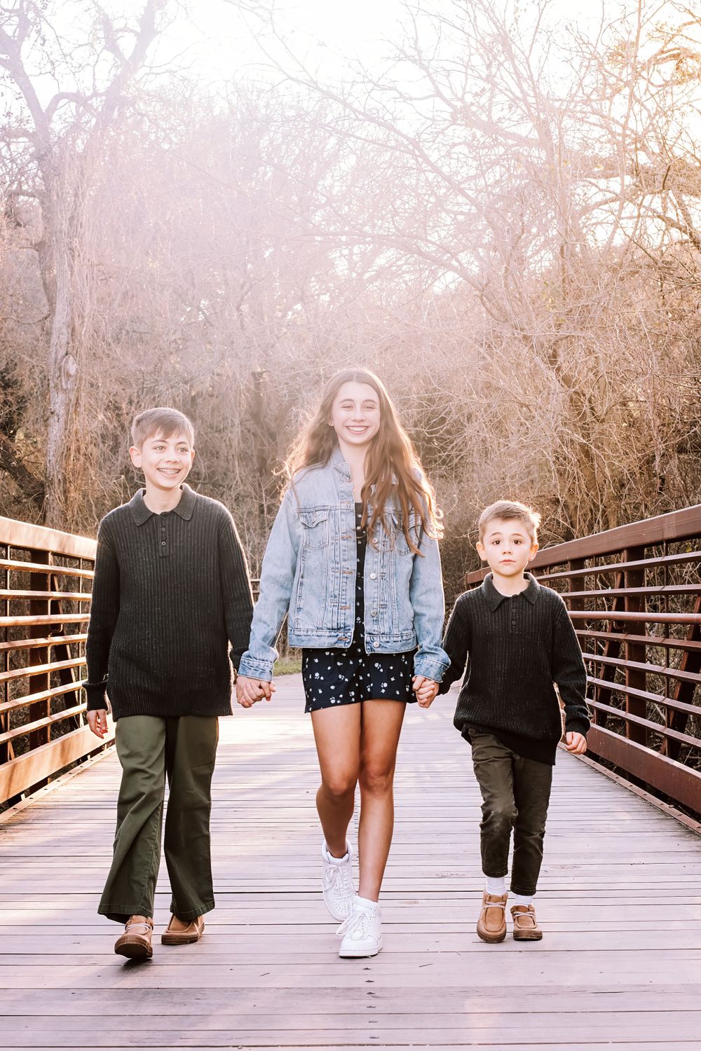 Three siblings walking hand in hand along a wooden bridge pathway on a misty morning.