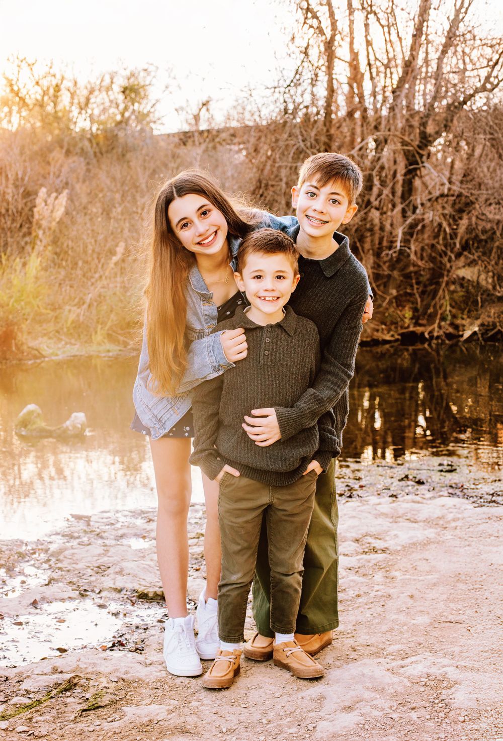 Three siblings share a happy moment together by a lake at sunset in warm autumn lighting.