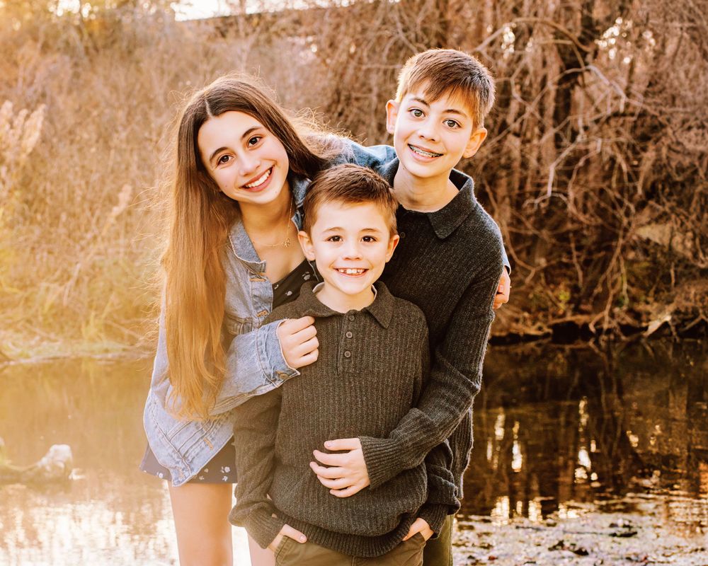 A happy family of three poses outdoors by a lake during golden hour in autumn while wearing cozy sweaters.