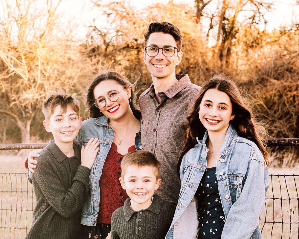 A group of five people wearing casual clothing smiles together outdoors during autumn with golden foliage in the background.