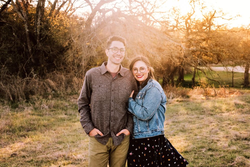 Two people pose together outdoors at sunset with autumn trees and golden light in the background.