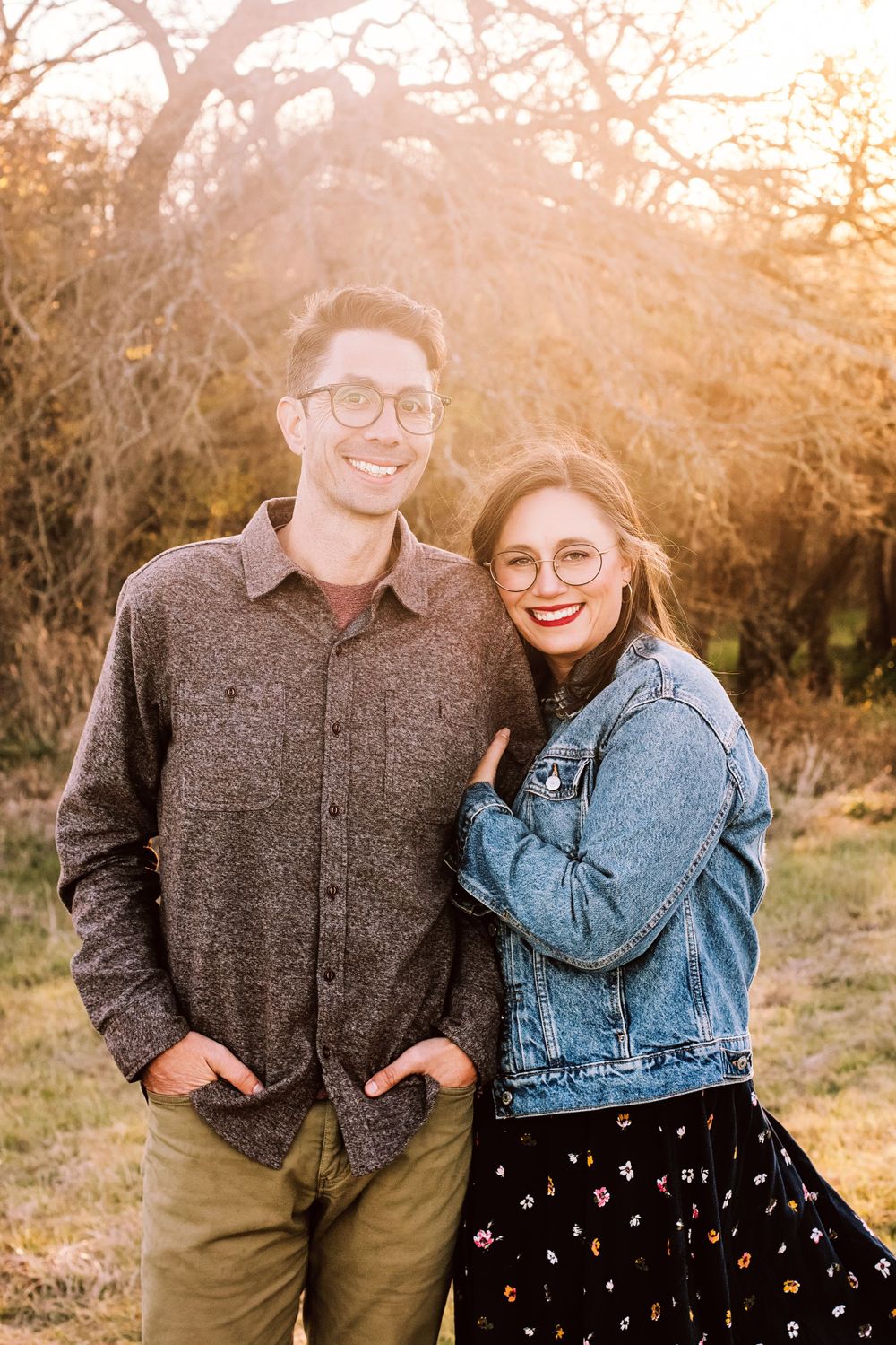 A couple wearing glasses poses together outdoors at sunset, with autumn trees in the background.