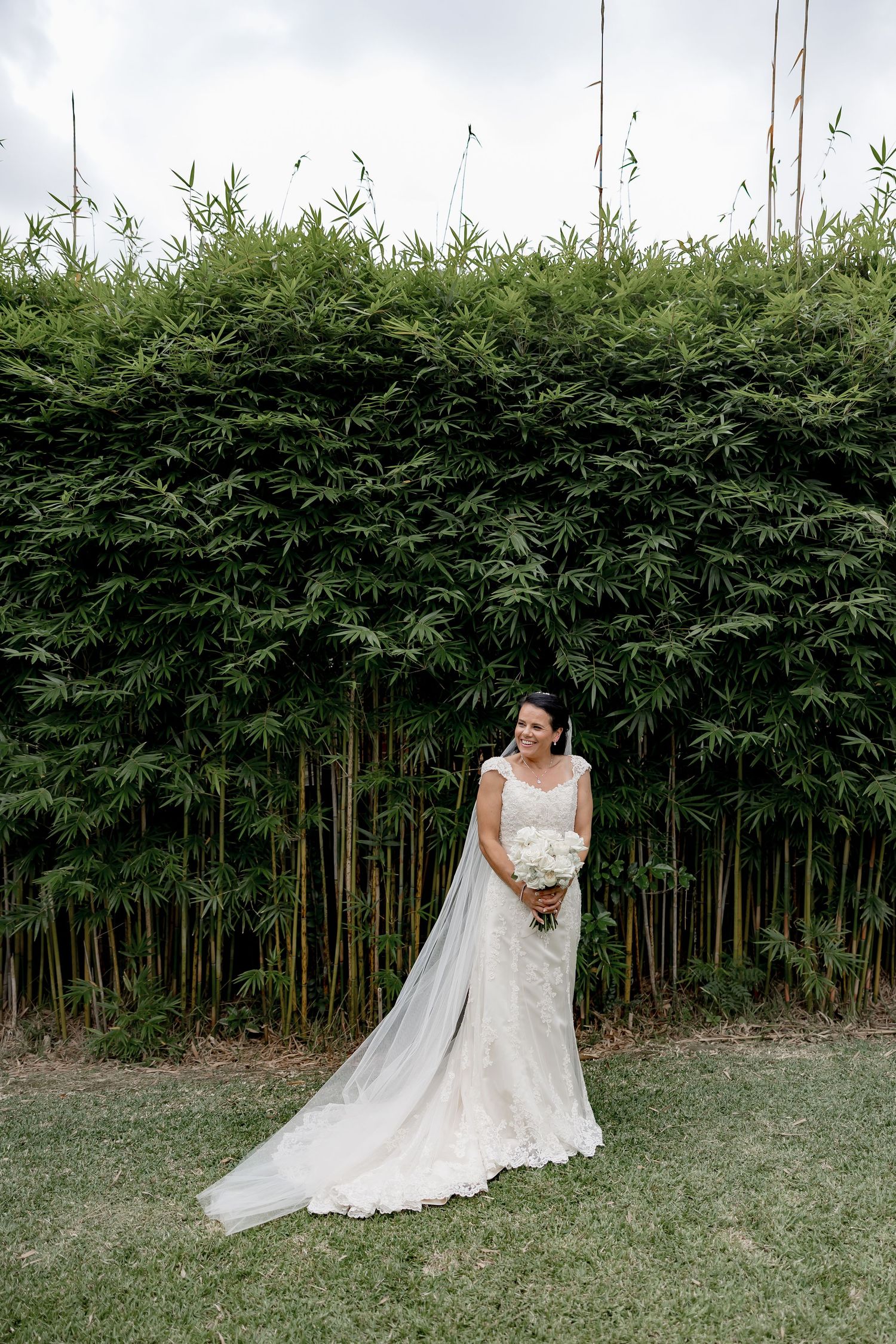 An elegant lace wedding dress stands out against a tall bamboo hedge backdrop in an outdoor garden setting.