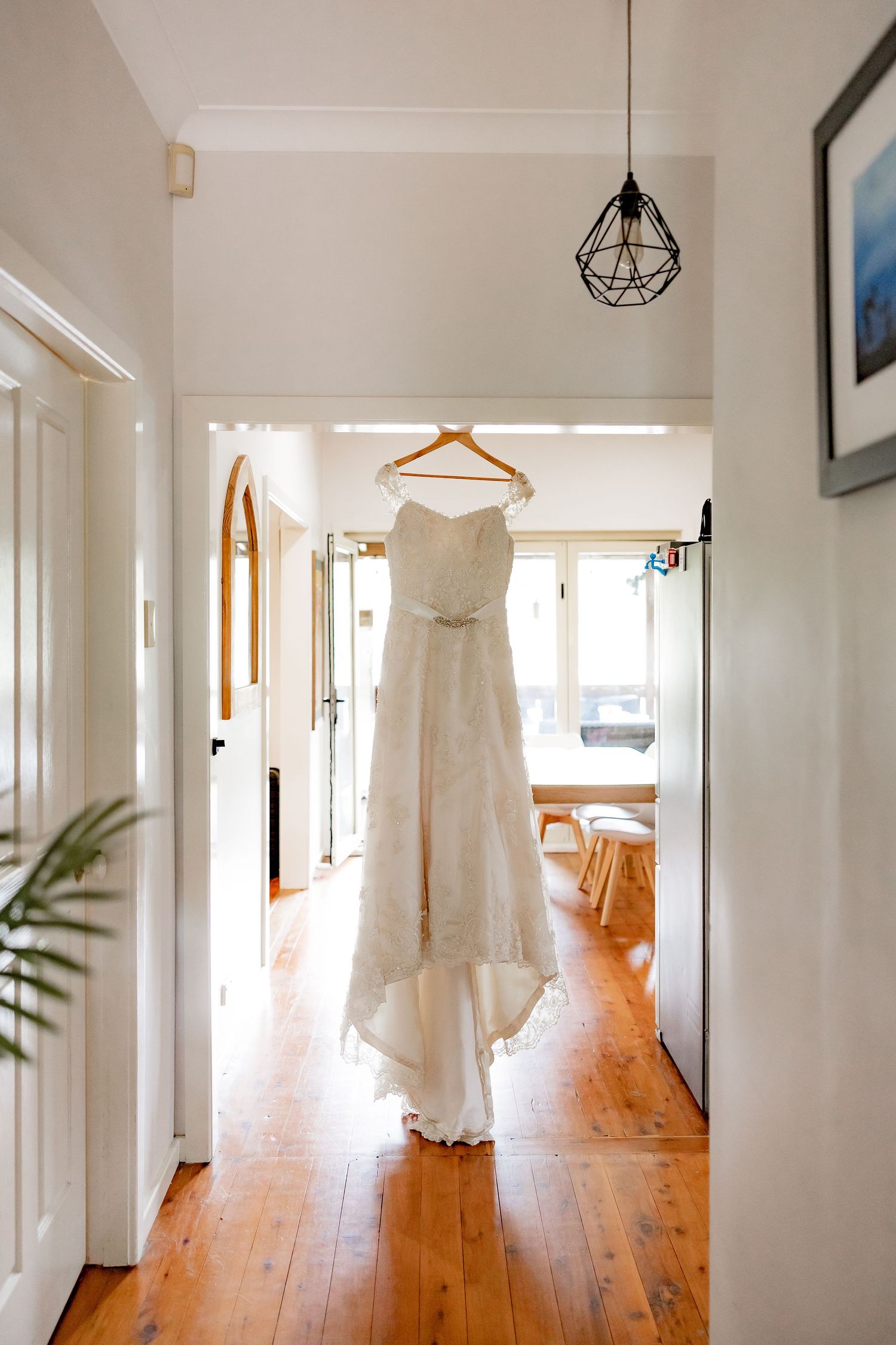 An elegant white lace wedding dress hangs from a wooden hanger in a hallway with hardwood floors and geometric pendant light.