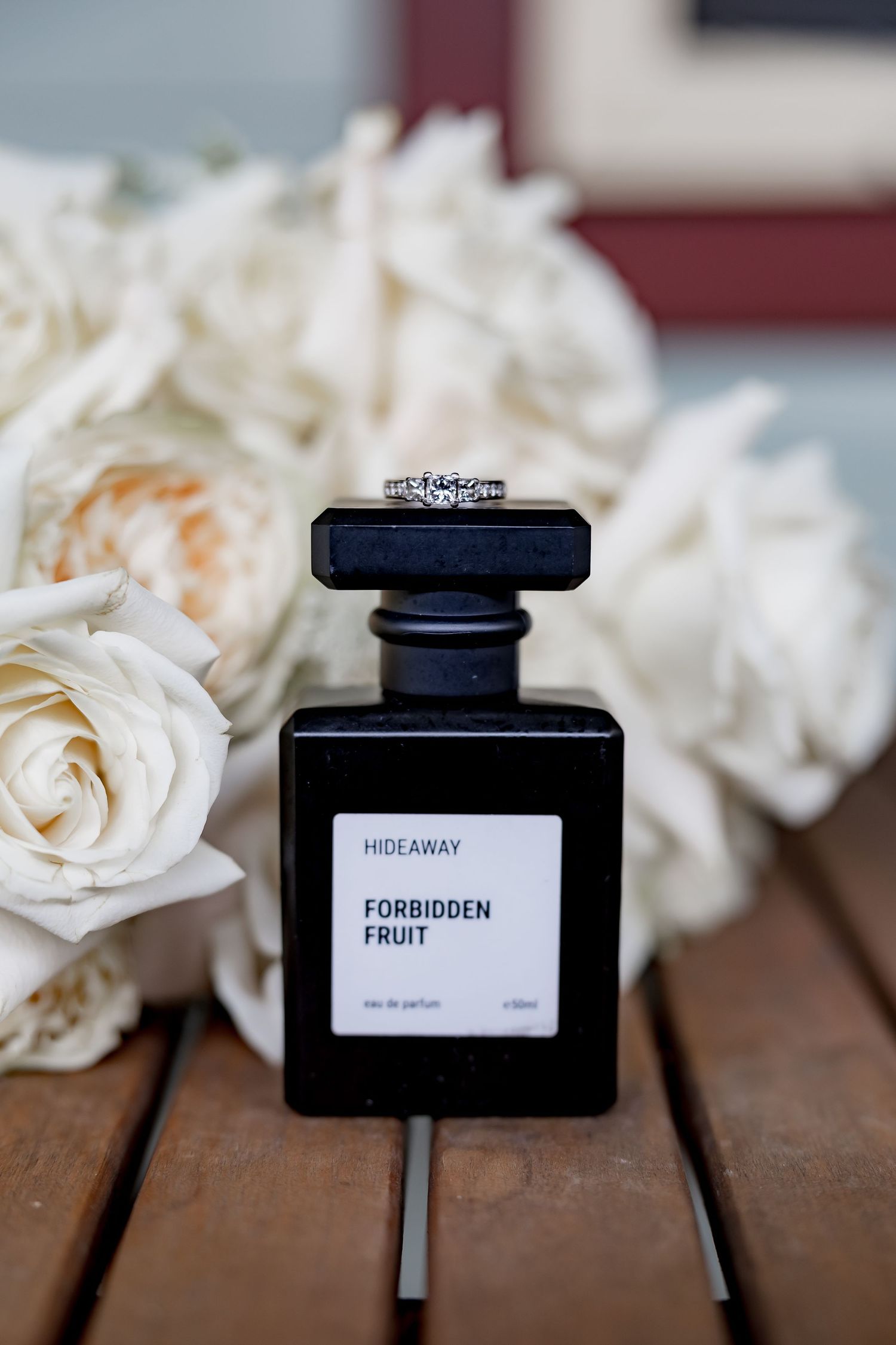 A diamond engagement ring sits atop a black Forbidden Fruit perfume bottle surrounded by white roses.