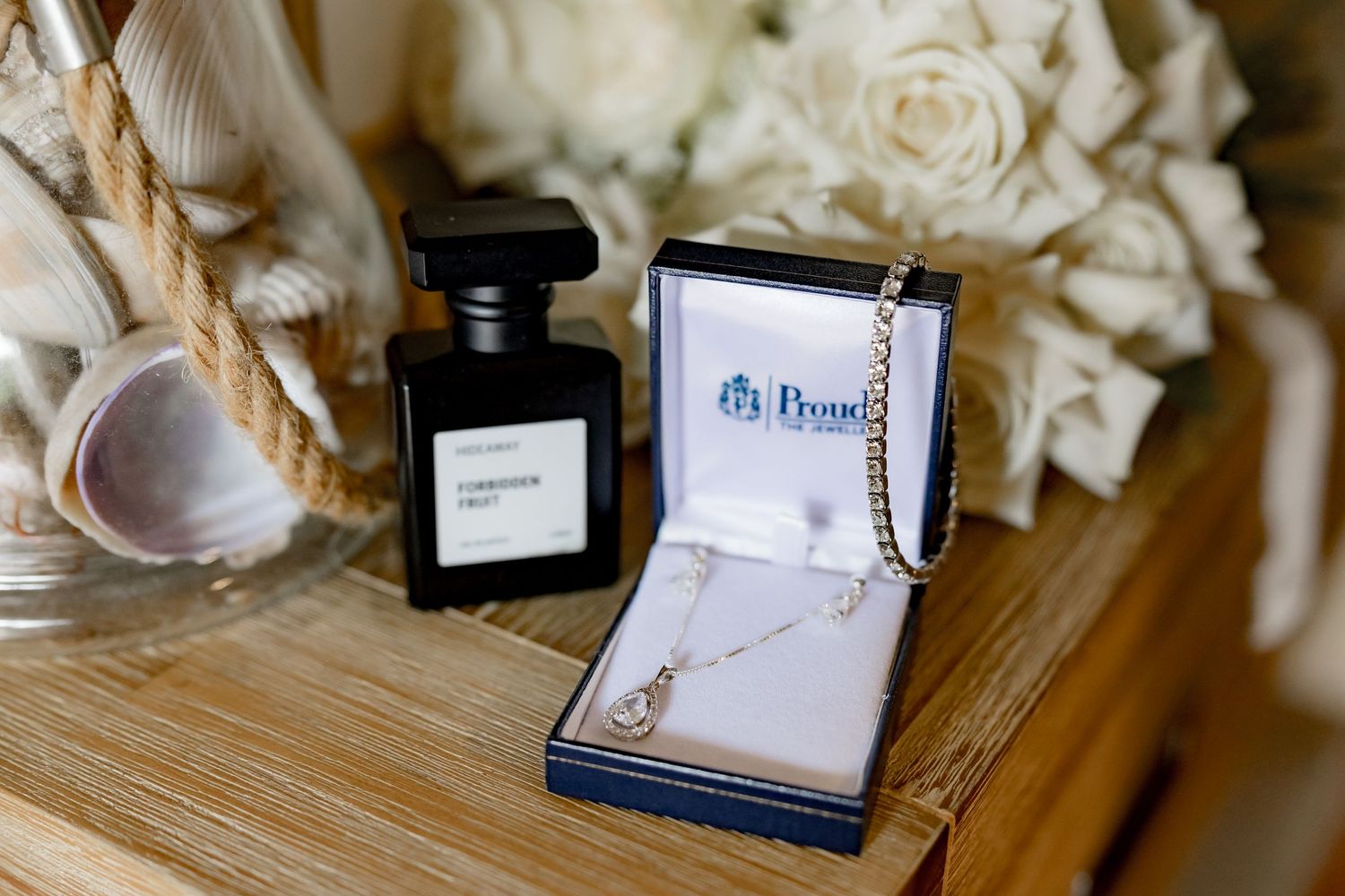 An elegant jewelry necklace box sits on a wooden surface with black perfume bottle and white roses in the background.