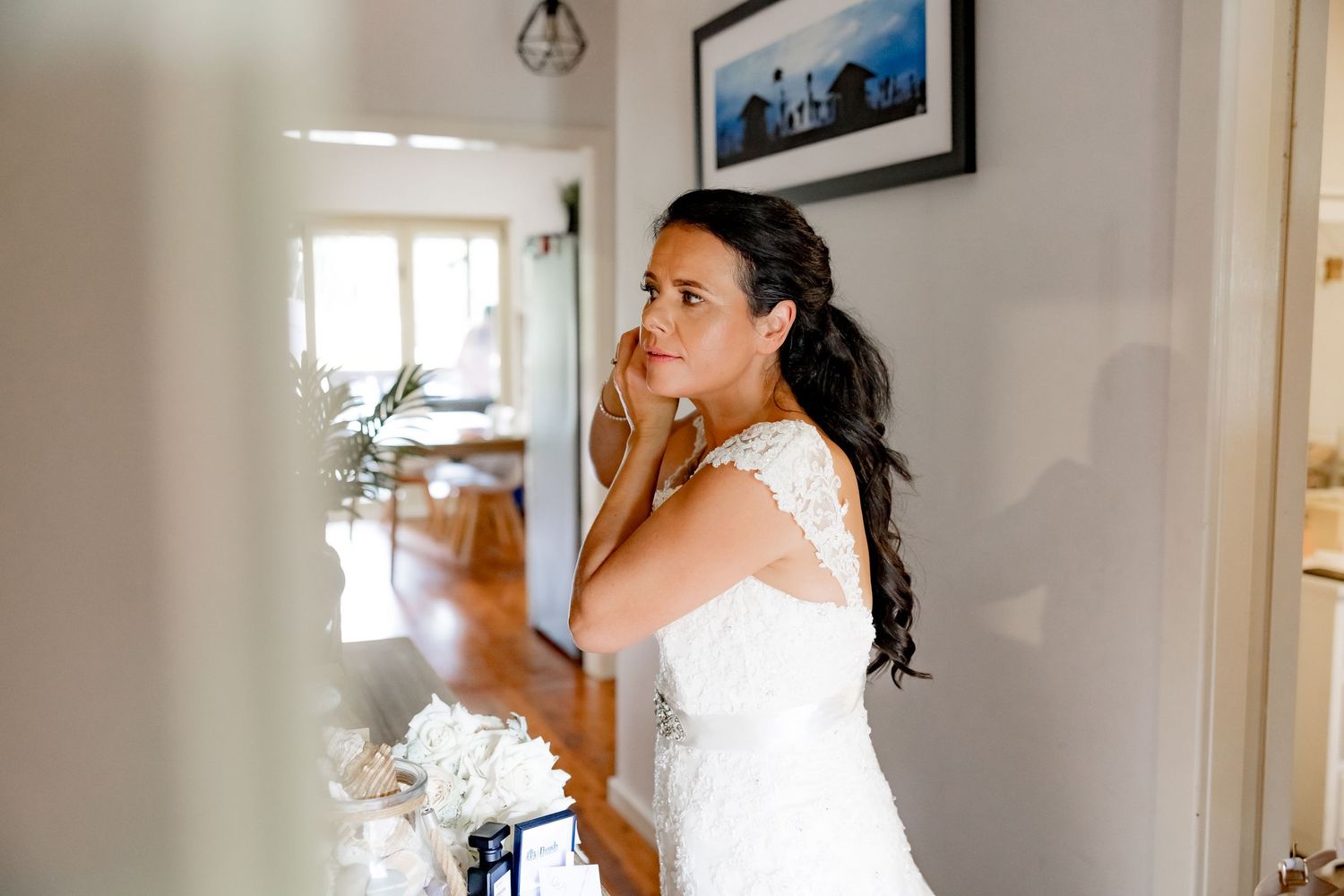 Someone in a white lace wedding dress stands by a window while putting on earrings in preparation for their big day.