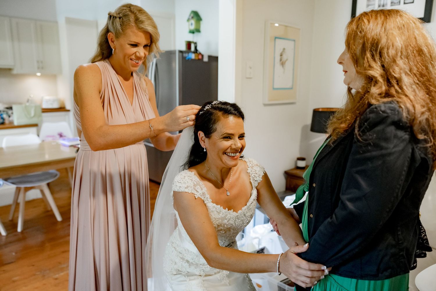 A bride in white dress gets ready for her wedding day with help from two attendants in a bright home setting.