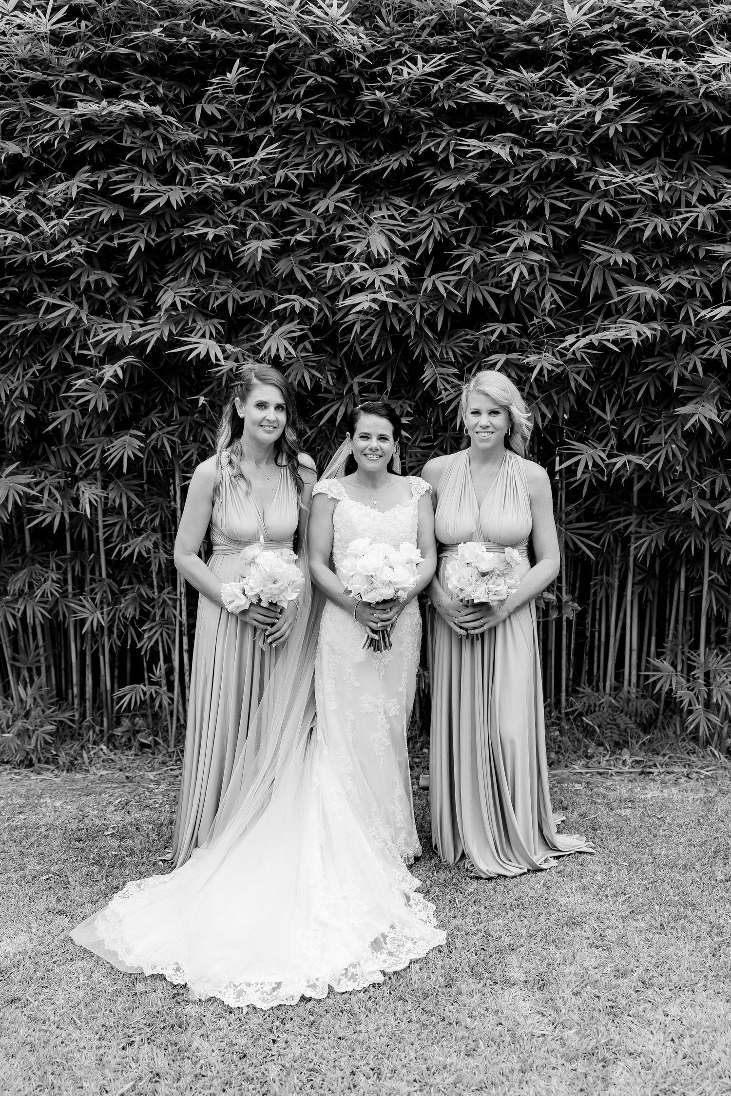 Three women in elegant gowns stand in front of a bamboo backdrop with bridal bouquets.