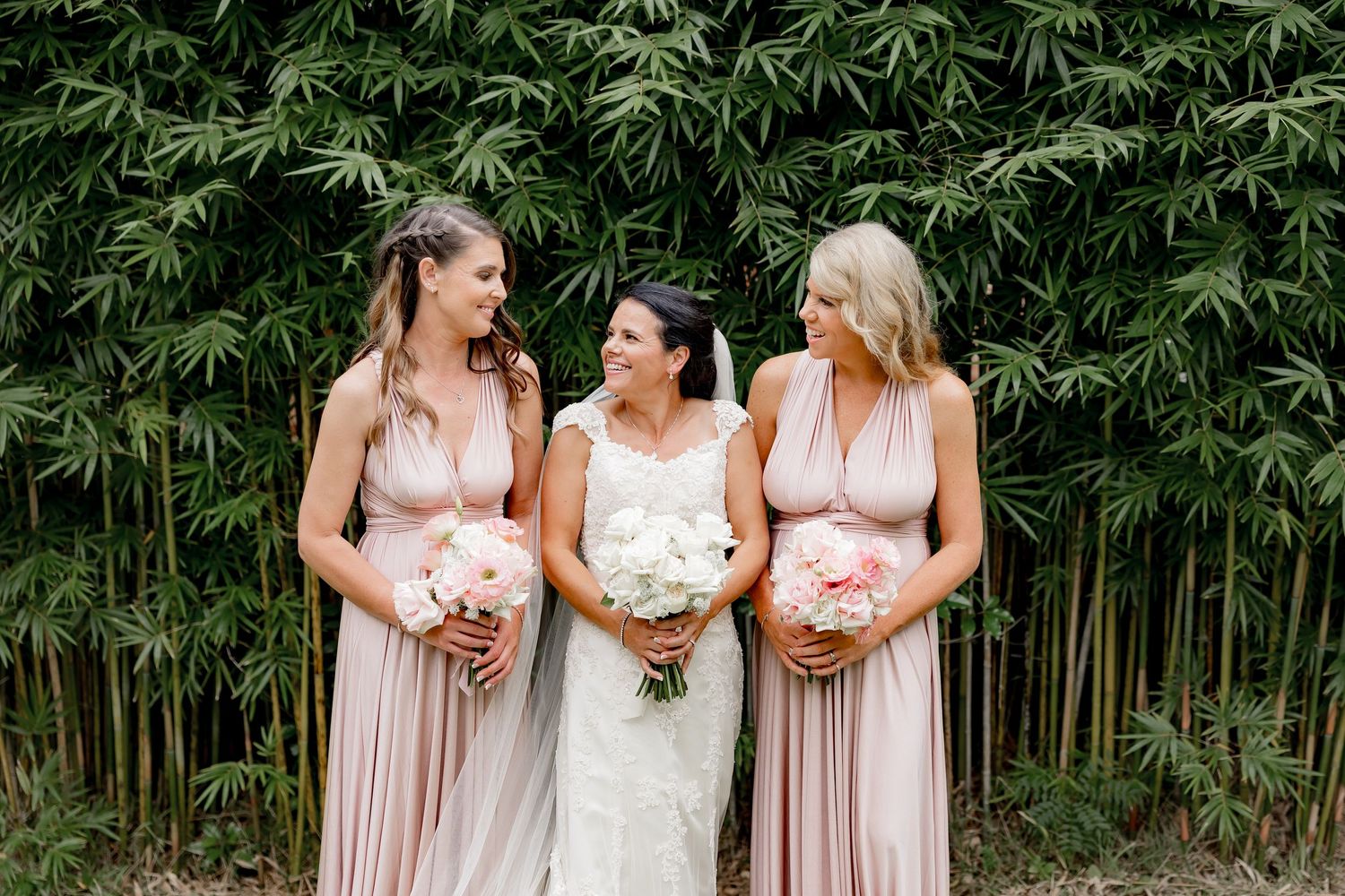 Three people wearing formal dresses pose together against a bamboo backdrop holding pink floral bouquets.