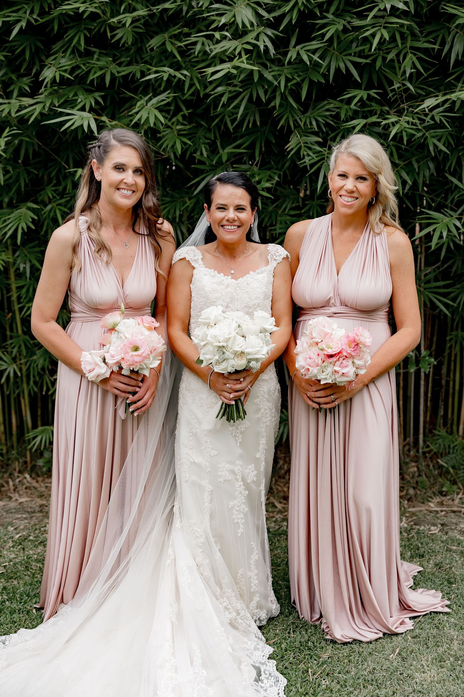Three women in pink and white gowns holding bouquets pose outdoors with bamboo backdrop at a wedding celebration.