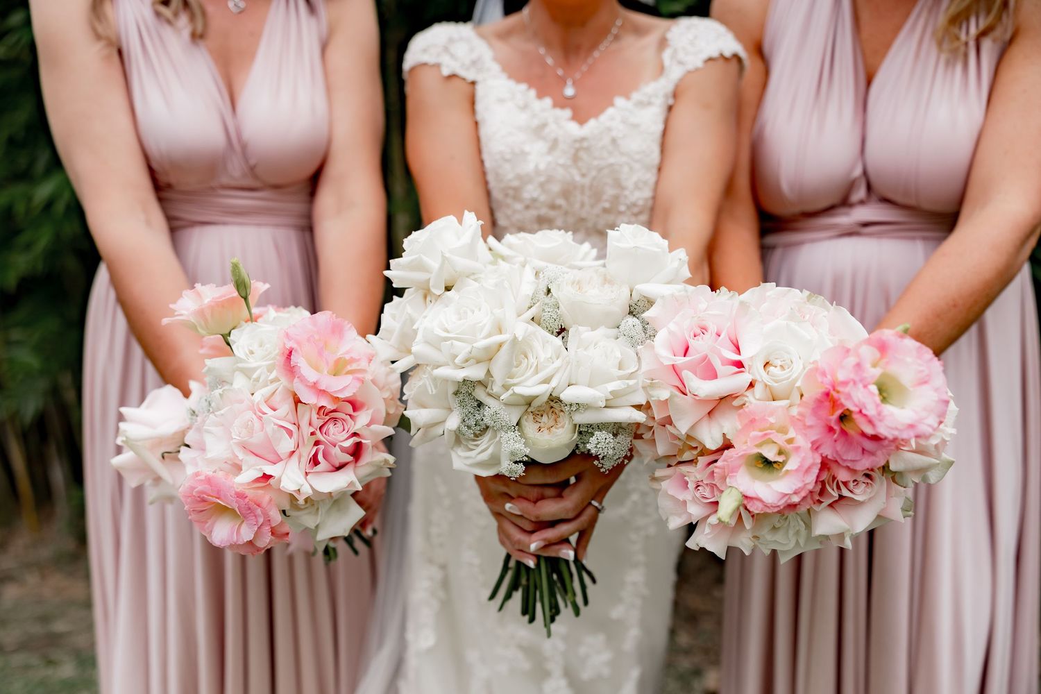 Three wedding bouquets featuring white and pink roses held against blush pink bridesmaid dresses at an outdoor ceremony.