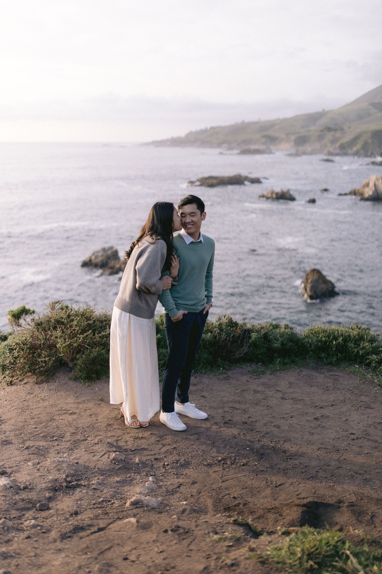 A couple shares a romantic moment on a cliff overlooking a rocky Pacific coast during sunset.