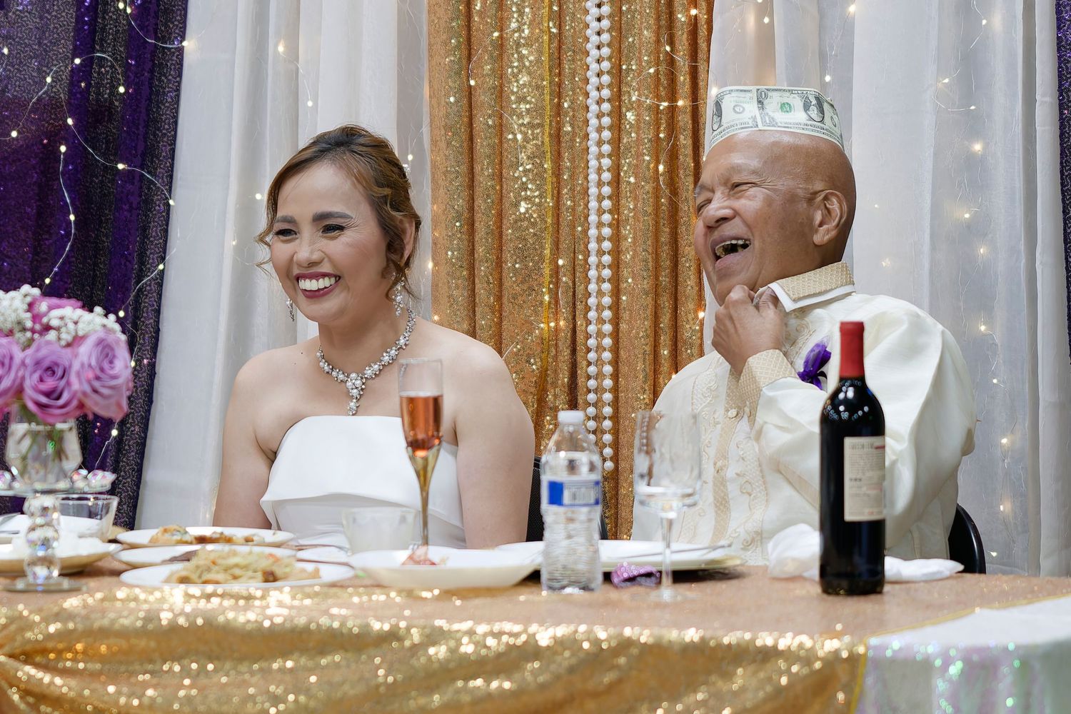 A couple in formal wedding attire sits together at a reception with purple and gold decorations in the background.
