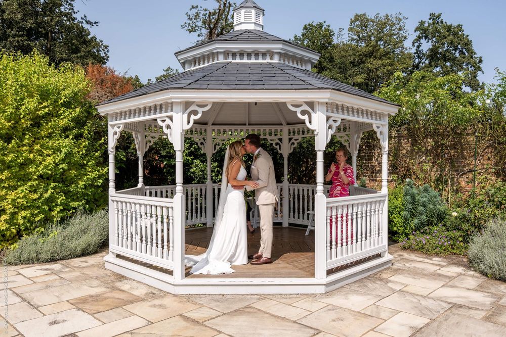 A elegant white gazebo in a garden setting with wedding ceremony participants gathered inside.
