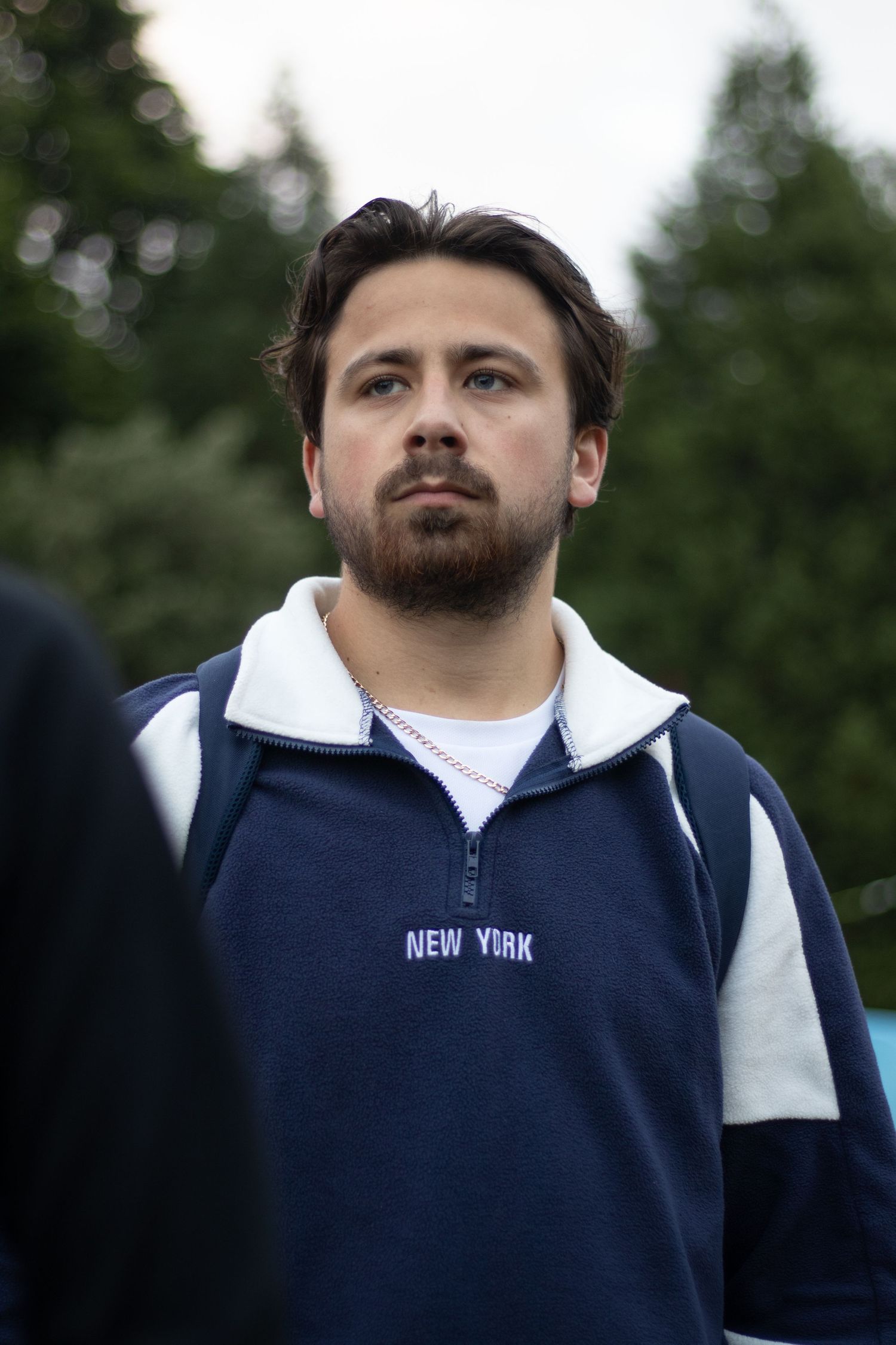 Person wearing navy and white New York sweatshirt stands outdoors against green foliage.