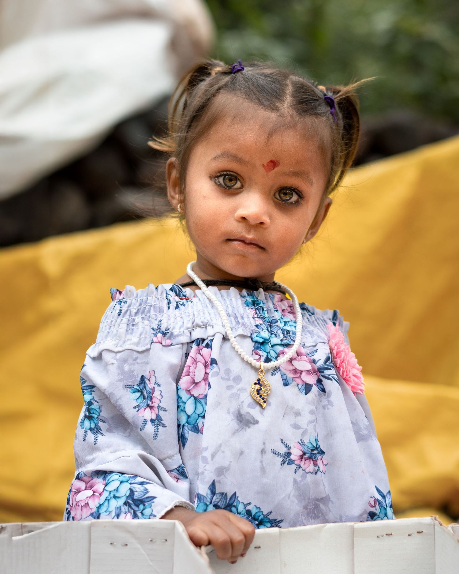 Young child in floral dress with pigtails looks at camera against yellow background.
