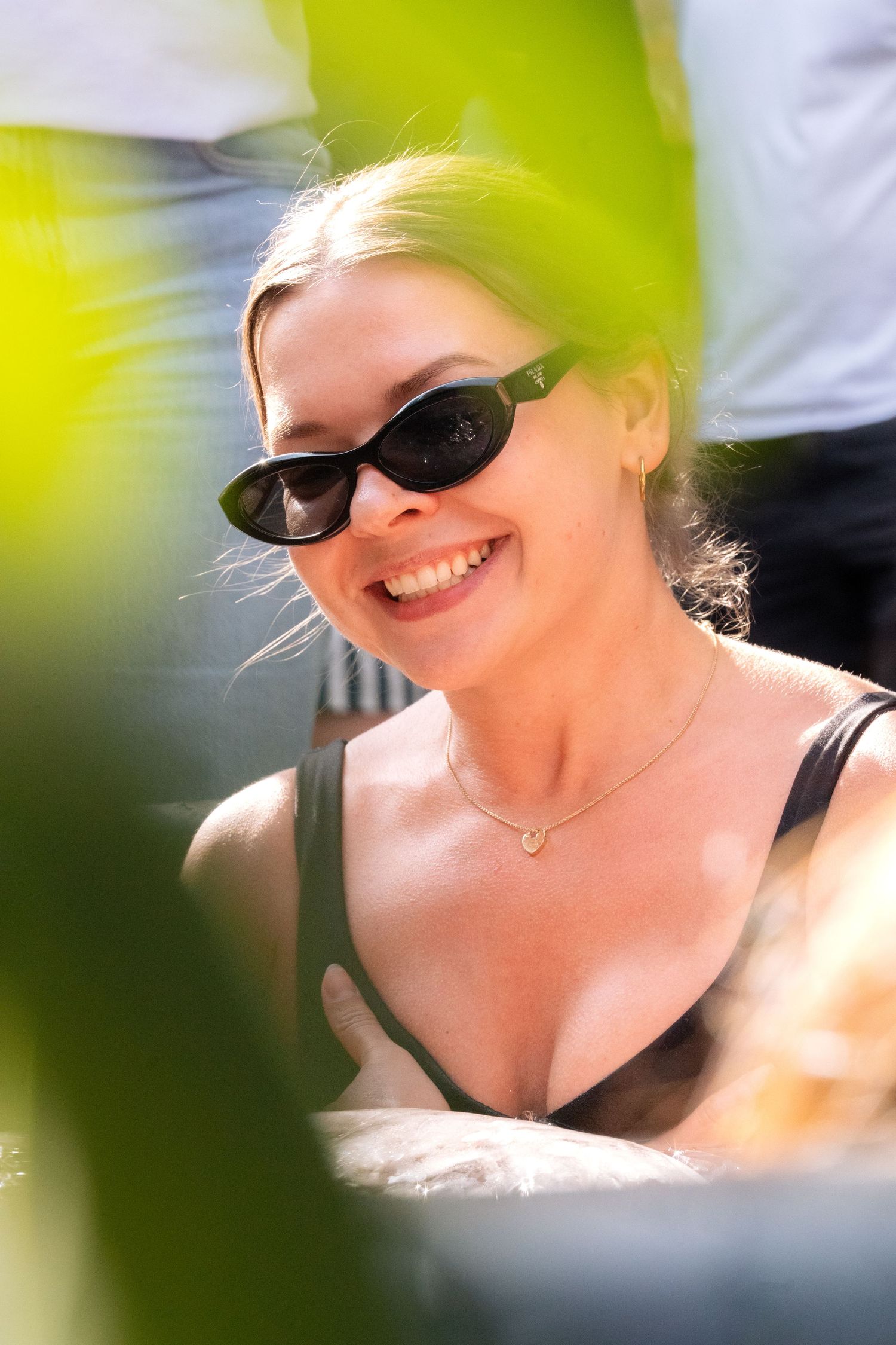 Person in black sunglasses and tank top smiles brightly while sitting outdoors on a sunny day.