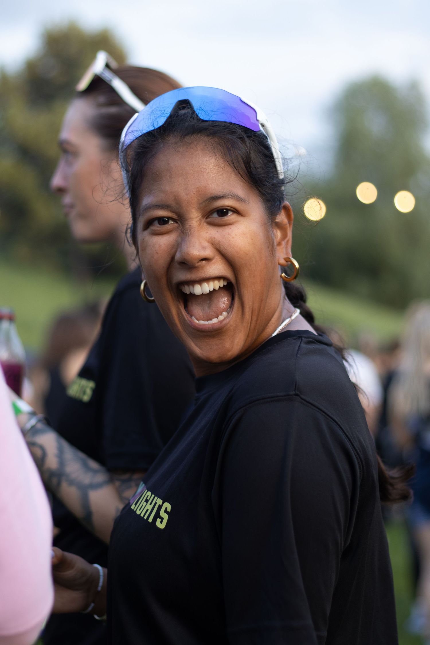 A person in sporty attire and sunglasses laughs joyfully during an outdoor event with twinkling lights in the background.