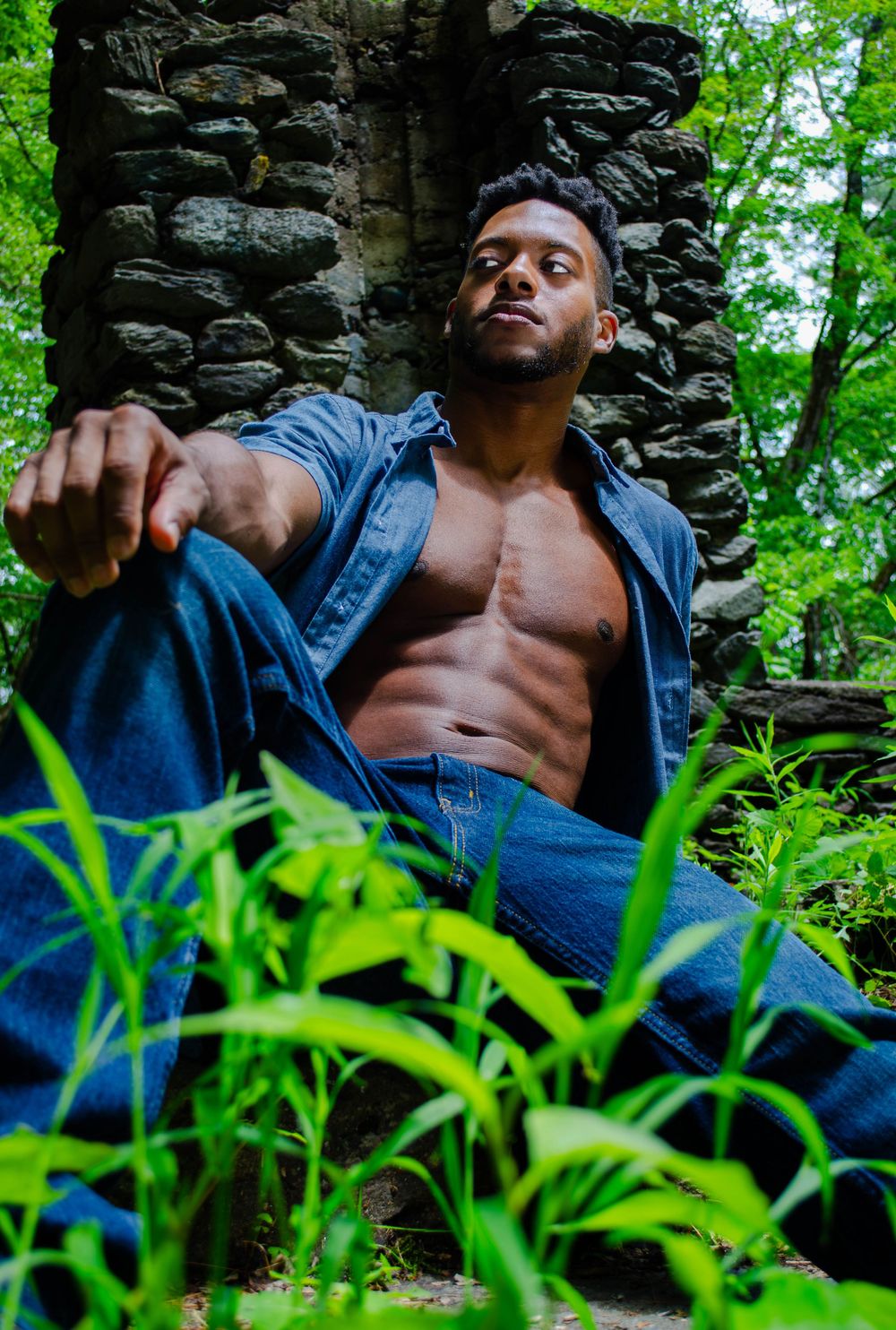 Person in denim jacket poses against stone wall surrounded by lush green foliage in artistic outdoor photoshoot.