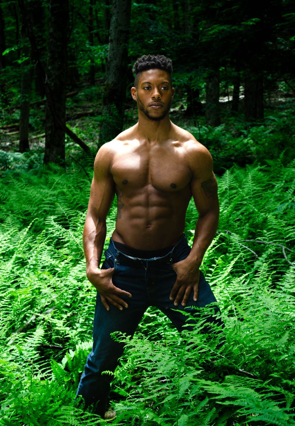 A shirtless person poses dramatically among lush green ferns in a forest setting.