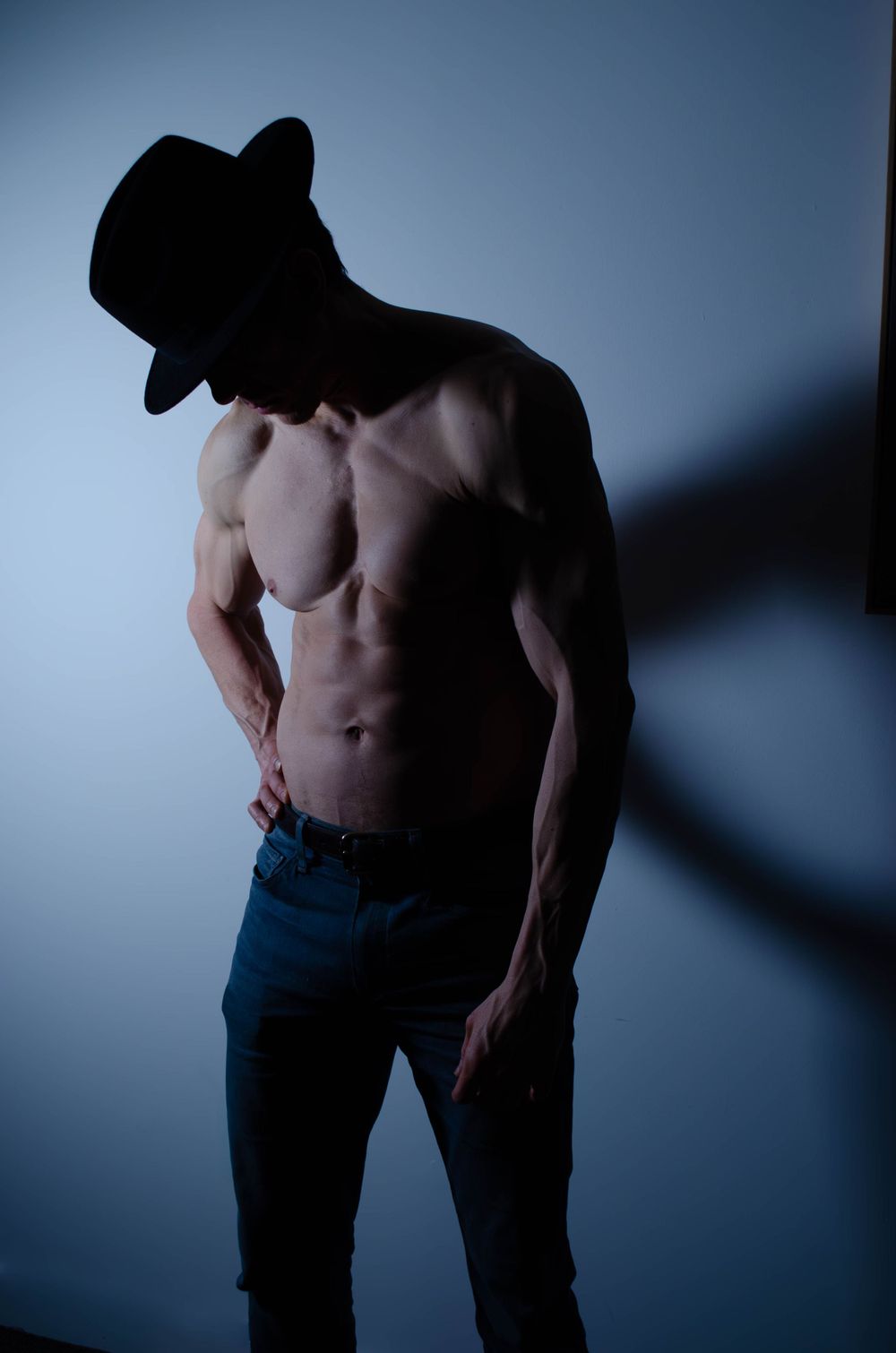 Men's Boudoir. Dudeoir. Artistic shadowy portrait of muscular torso in hat against blue background.