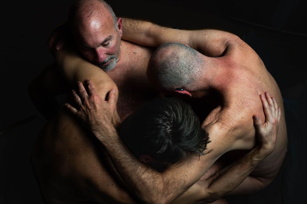 Two figures embrace intimately in an artistic dark-lit studio photograph against black background.