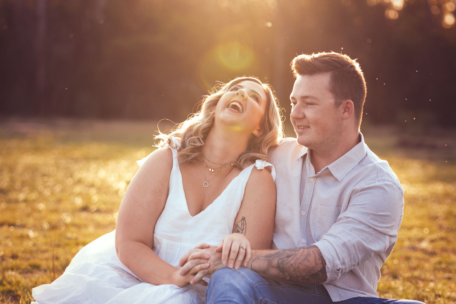 A couple is sitting on the grass in a sunlit field. The woman is laughing and leaning towards the man, who is smiling and holding her hand.